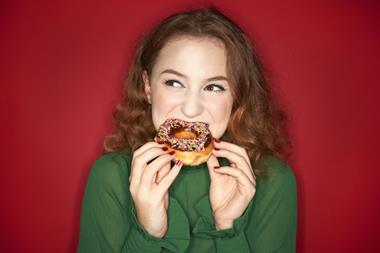 Credit Tara Moore via GettyImages_Young woman eating iced donut on red background