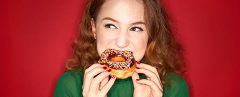 Credit Tara Moore via GettyImages_Young woman eating iced donut on red background