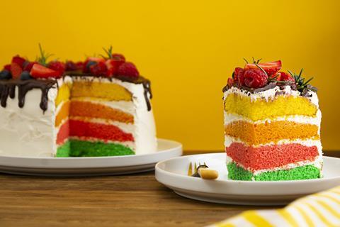 Slice of fruit-topped rainbow cake on a plate with rest of cake on the background.